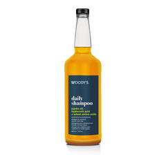 A bottle of Woody's Daily Shampoo featuring a clear glass bottle filled with golden-yellow liquid, a black cap, and a dark navy blue label with white text.