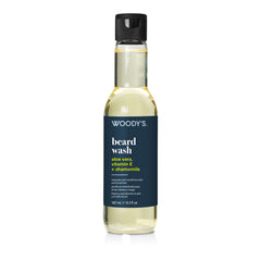 A bottle of Woody's Beard Wash with a dark navy blue label and a black cap, containing a pale yellow liquid, shown on a white background.