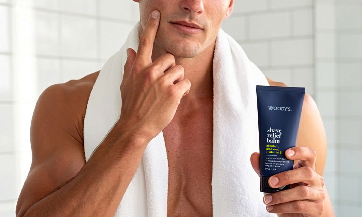 A man with a white towel over his shoulders touching his smooth, freshly shaved cheek while holding a tube of Woody's Shave Relief Balm.