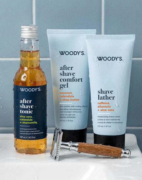 Woody's men's grooming set with a small bottle of after shave tonic, a tube of after shave comfort gel, and a tube of shave lather, alongside a folding razor on a white sink against a blue-gray tiled wall.
